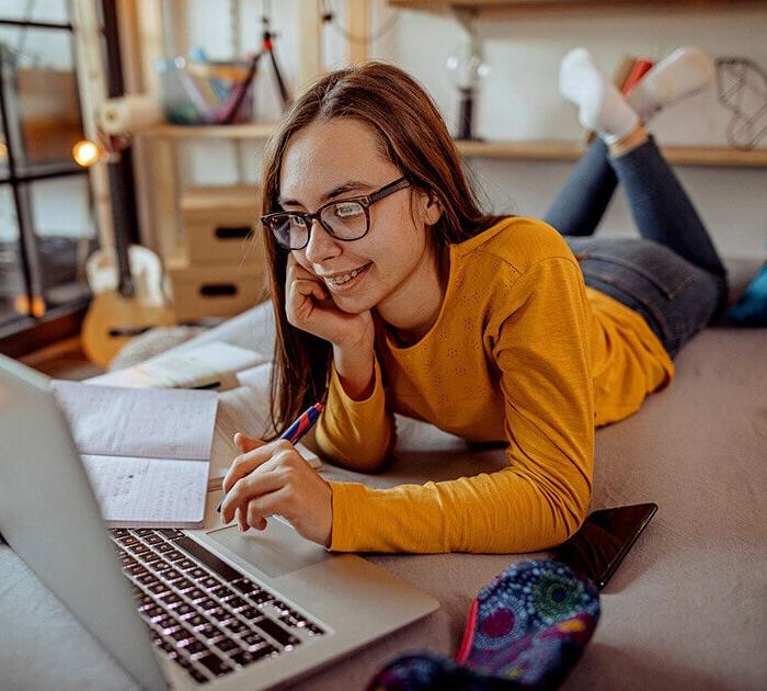 A teenage girl laying across her bed taking notes during her online class