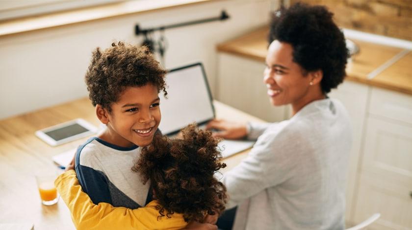 A mother is sitting at a computer while her son and daughter hug eachother